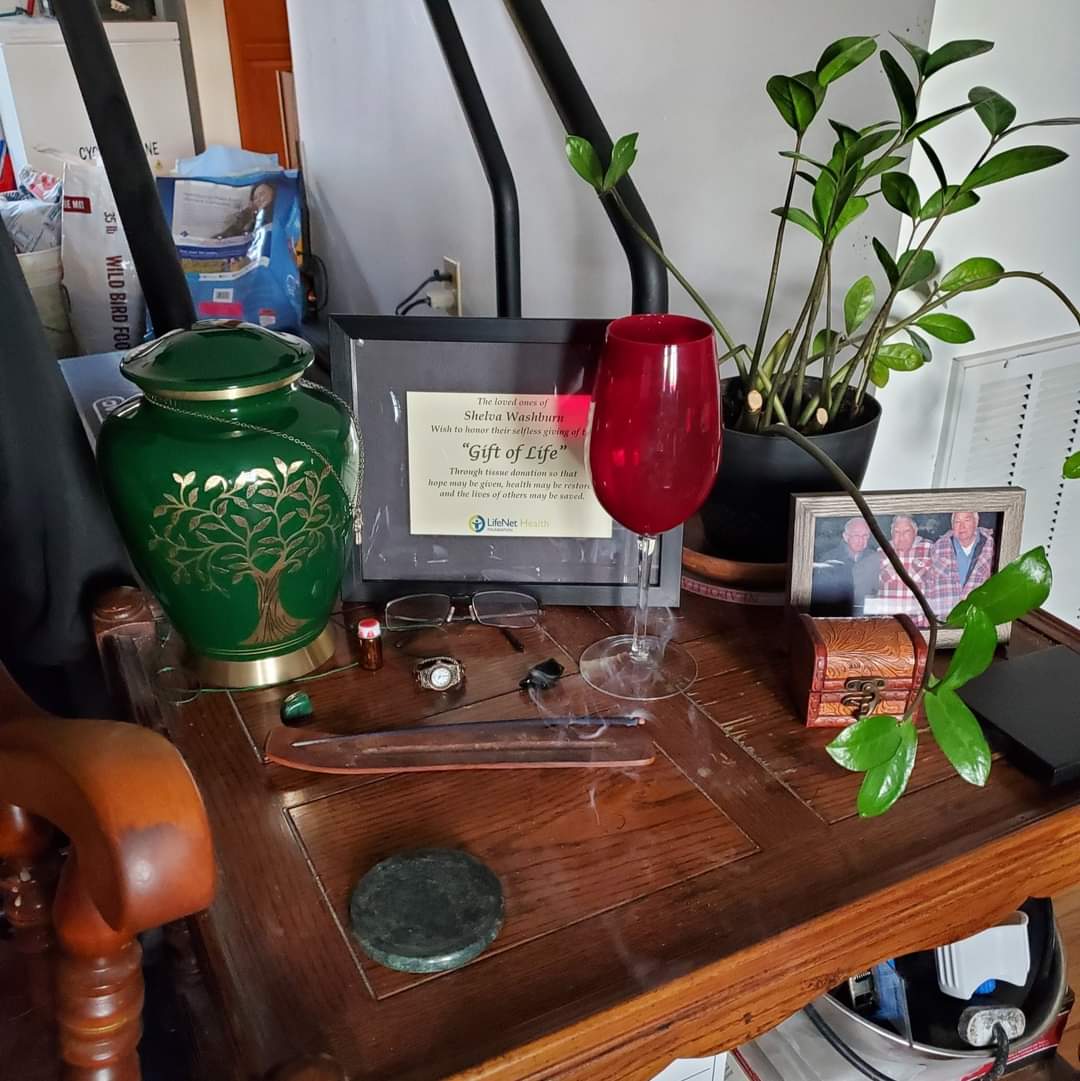 Shelva's urn sitting on a table with a red wine glass with water, an incense, glasses, a watch, a certificate showing she was an organ donor, a plant, a picture showing her dad, and a small amount of her dad's cremains.